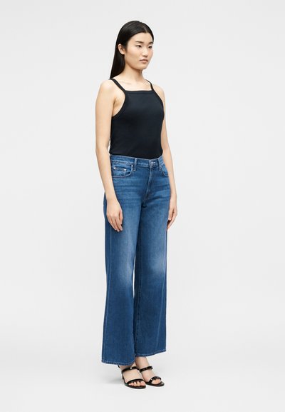 Γυναίκα στέκεται φορώντας ένα μαύρο αμάνικο τοπ, μπλε φαρδιά τζιν και μαύρα σανδάλια με λουράκια σε απλό ανοιχτό φόντο.