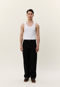 Νεαρός άνδρας στέκεται όρθιος φορώντας λευκό αμάνικο τοπ, μαύρο παντελόνι φαρδύ στο τελείωμα και καφέ παπούτσια σε φωτεινό φόντο.