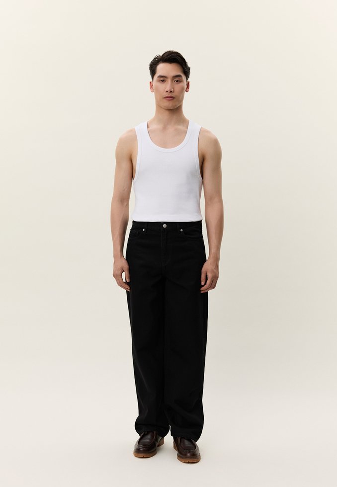 Νεαρός άνδρας στέκεται όρθιος φορώντας λευκό αμάνικο τοπ, μαύρο παντελόνι φαρδύ στο τελείωμα και καφέ παπούτσια σε φωτεινό φόντο.