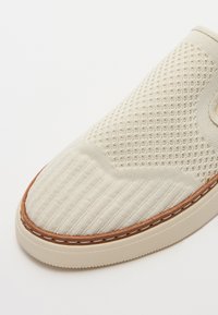 Hele beež slip-on king, millel on võrk ülemine osa, millel on tekstuursed mustrid, kummist tallad ja ääres õmblemise detailid.