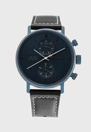 Montre-bracelet ronde noire Hugo avec deux petits cadrans indiquant le jour et la date, aiguilles argentées, bracelet en cuir noir avec coutures blanches.