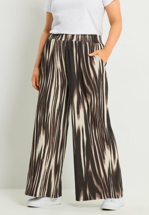 Osoba ubrana w spodnie o szerokich nogawkach z czarno-beżowymi abstrakcyjnymi paskami, białe trampki i białą koszulę z krótkim rękawem, stoi z jedną ręką w kieszeni.