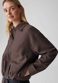 Braune Jacke mit Reißverschluss, Kragen, oversized Schnitt und entspannter Passform. Hergestellt aus strukturiertem Stoff mit Seitentaschen und elastischem Saum.