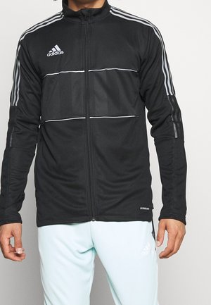 Homme portant une veste zippée Adidas noire avec des bandes blanches sur les manches et un pantalon Adidas clair sur fond blanc.