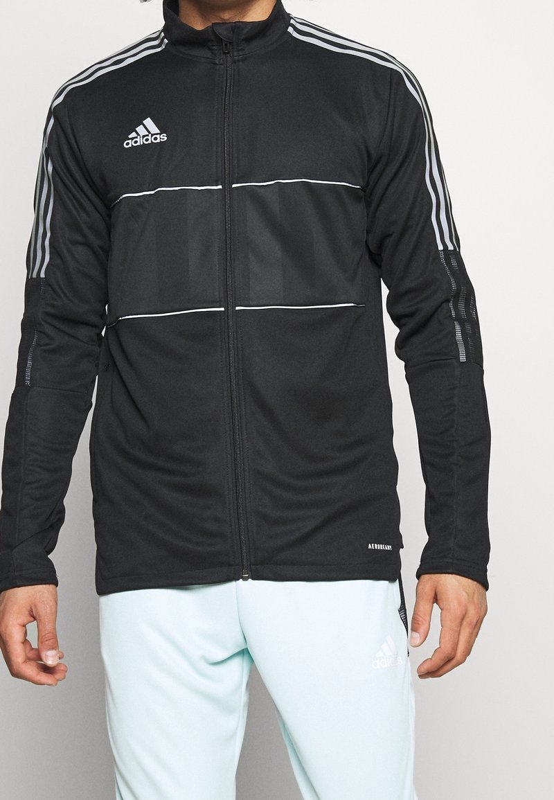 Homme portant une veste zippée Adidas noire avec des bandes blanches sur les manches et un pantalon Adidas clair sur fond blanc.