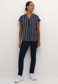 Camisa de rayas en azul marino con mangas cortas y tapeta de botones, combinada con jeans ajustados de un azul oscuro y zapatillas blancas sobre un fondo claro.