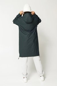 Pitkä tummanvihreä vedenpitävä takki, jossa on huppu, vetoketjullinen etuosa ja säädettävät nyörit, yhdistettynä harmaisiin collegehousuihin ja valkoisiin lenkkareihin.