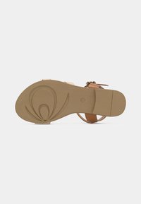 Κάτω όψη σανδαλιού που παρουσιάζει μια λεία, μπεζ σόλα με λουλουδένιο σχέδιο και καφέ λουριά στερεωμένα με αγκράφα σε χρυσό τόνο.