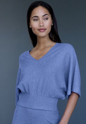 Frau mit langen dunklen Haaren, die ein blaues, geripptes V-Ausschnitt-Top mit kurzen Ärmeln vor einem schlichten dunkelgrauen Hintergrund trägt.