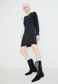 Vestido de malha cinza com mangas longas e gola alta; combinado com botas de tornozelo de couro pretas. Um gorro rosa completa o visual.