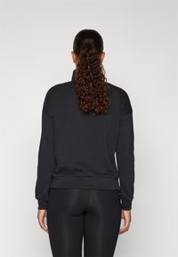 Kobieta z kręconym kucykiem, ubrana w czarną bluzkę z długim rękawem i czarne legginsy, stojąca na jednolitym jasnoszarym tle.