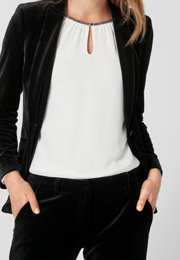 Musta samettitakki valkoisen, avokauluksisen puseron päällä, jossa on koristeellinen hopeinen reunus. Samettiset mustat housut täydentävät asun.