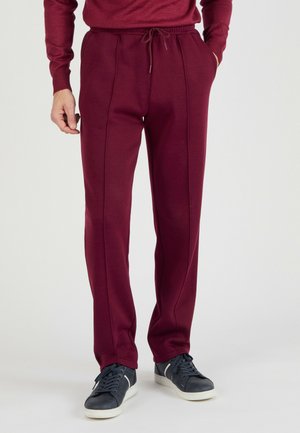Man draagt bordeauxrode joggingbroek met voorzakken en zwarte sneakers met witte zolen, staand op een effen lichte achtergrond.
