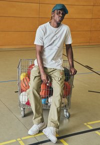 Fehér póló grafikus logóval, olajzöld nadrág "NBA" emblémával és fehér tornacipő. Egy drótkosárban különböző kosárlabdák találhatók.