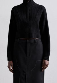 Pull court noir en maille avec un col montant et une texture côtelée, associé à une jupe noire avec des détails de couture contrastants et un bouton.