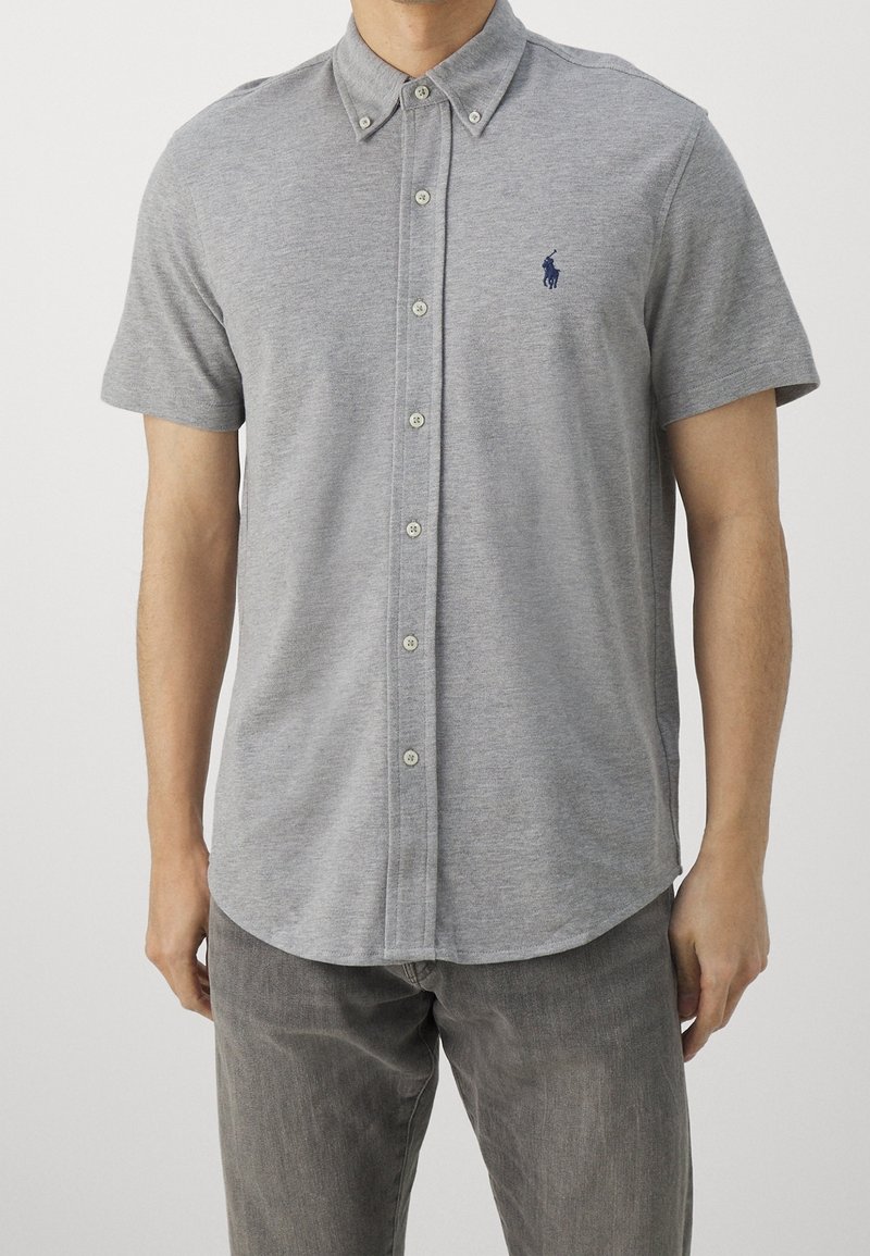 Camicia grigia a maniche corte con bottoni, realizzata in un tessuto leggero, con colletto classico e un piccolo logo ricamato sul petto.