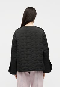 Άτομο με μακριά σκούρα μαλλιά που φοράει μαύρο καπιτονέ μπουφάν με κυματιστά σχέδια, όρθιο με το σώμα στραμμένο προς τα πίσω, σε συνδυασμό με ανοιχτό μωβ παντελόνι.