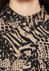 Blouse met kraag van beige stof met zwarte abstracte patronen en gouden knoopaccenten. Getextureerd, lichtgewicht materiaal.