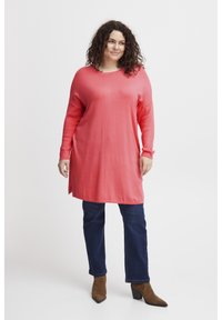 Tunika a maniche lunghe in tessuto a maglia corallo con scollatura rotonda e spacchi laterali, abbinata a jeans blu scuro e stivaletti con tacco marroni.