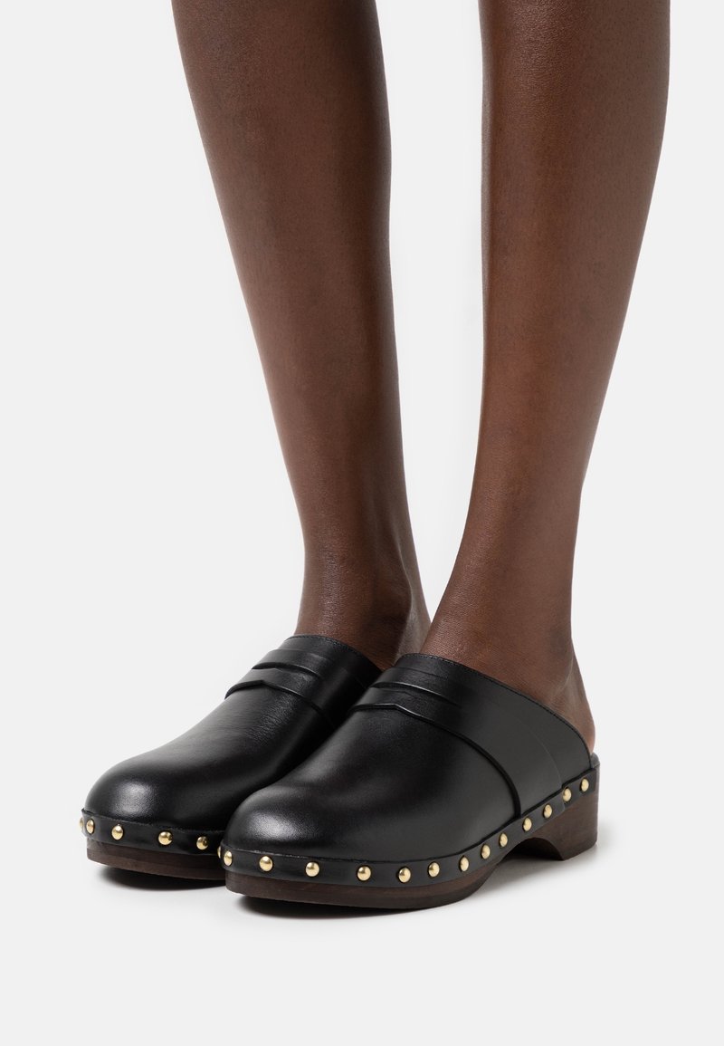 Sabots en cuir noir avec un bout rond, un petit talon en bois, deux bandes surélevées sur le dessus et des clous dorés le long de la semelle.