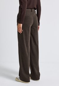 Bruna, högmidjade byxor med ett texturerat mönster, som har sidofickor och en avslappnad passform, ihop med vita och grå sneakers.