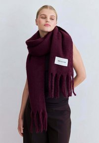 Bordeaux wolmix sjaal met franjes en een wit label met daarop "ARCTIC FOX," elegant om de nek gedrapeerd. Zachte textuur.