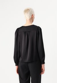 Blusa de satén negro con mangas abullonadas, escote redondo y detalles fruncidos en la parte posterior. Textura suave y ajuste entallado.