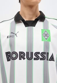 Άνδρας που φοράει λευκό ποδοσφαιρικό πουκάμισο με μαύρες και πράσινες κάθετες ρίγες, μαύρο γιακά, λογότυπο της Μπορούσια, λογότυπο Puma και το κείμενο "BORUSSIA".