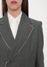 Grå dobbelspent blazer med synlig hvit søm. Har notched revers, en rød aksent og knappehull. Båret over en hvit skjorte.