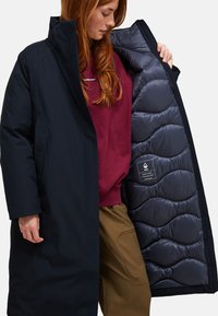 Persoon in een lange zwarte gewatteerde jas, waarbij de gevoerde binnenvoering met een label zichtbaar is, gekleed in een bordeauxrood topje en een bruine broek.