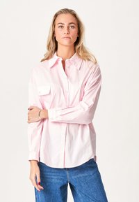 Ung kvinde med blond hår iført en lys pink button-up skjorte og blå jeans, stående med armene korslagt mod en enkel hvid baggrund.