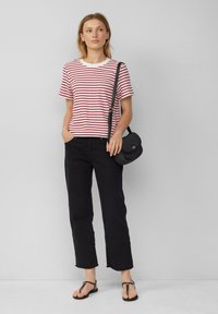 T-shirt rayé rouge et blanc, pantalons larges noirs et sandales plates. Le mannequin tient un sac à bandoulière noir, se tenant devant un fond neutre.