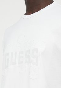 T-shirt bianca in cotone con logo "GUESS" in rilievo e motivi decorativi, dotata di scollo rotondo e maniche corte.