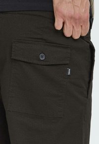 Primer plano de unos pantalones de tela oscura con un bolsillo abotonado y una pequeña etiqueta de marca, destacando la textura y los detalles. Una mano agarra la cinturilla.