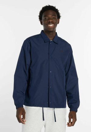 Junger Mann lächelt, trägt eine marineblaue Jacke mit Knöpfen und elastischen Bündchen sowie hellgraue Hosen vor weißem Hintergrund.