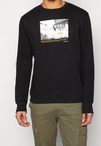 Felpa nera con stampa grafica e testo bianco e giallo. Polsini e fondo a coste. Tessuto morbido e strutturato. Indossata con pantaloni cargo color oliva.