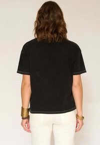 Une personne avec des cheveux bruns ondulés porte une chemise noire à manches courtes, un pantalon blanc et plusieurs bracelets en or, vue de dos.