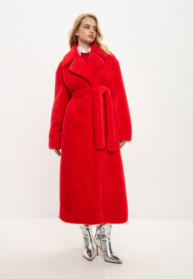 Abrigo de piel sintética roja con cuello chal, cinturón a la cintura y bolsillos laterales, combinado con botas de tacón alto metálicas. Textura suave.