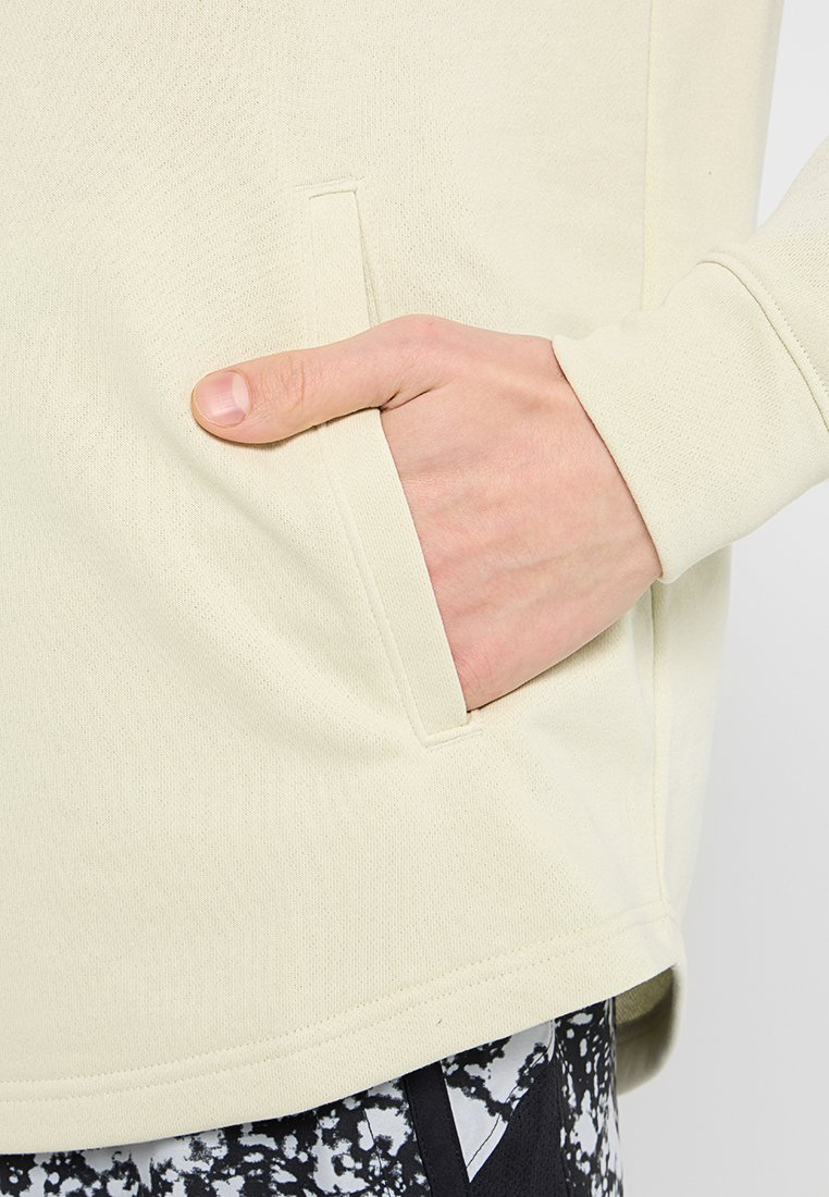 Pullover beige chiaro con tessuto strutturato, dotato di tasca laterale e design a vestibilità rilassata, abbinato a leggings neri con fantasia.