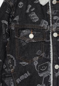 Jeansjacka i mörkgrått med grå grafiska mönster, fleece-fodrad krage, kontrasterande orange sömmar och silverfärgad knappdekoration.