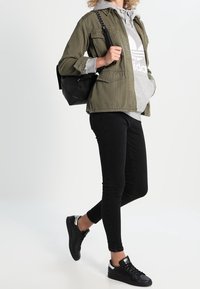 Olivově zelená bunda, šedá mikina s bílým logem, černé skinny džíny a černé tenisky. Nese černou tašku přes rameno.