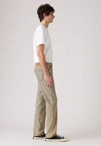 Joven de pie de perfil, vistiendo una camiseta blanca, jeans de color beige claro, zapatillas negras y un anillo de plata en su mano izquierda.