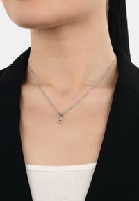 Kvinna som bär ett silver halsband med en liten röd ädelsten, kl�ädd i en svart blazer och vit topp.