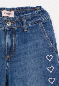 Μπλε τζιν παντελόνι με ελαστική μέση, ασημένιο κουμπί, κίτρινη ραφή και τρεις λευκές κεντητές καρδιές στο δεξί μηρό.
