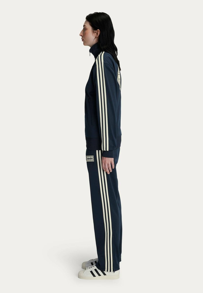Navy tracksuit with white stripes, made of smooth fabric. Features zippered pockets and an emblem on the back. Paired with white sneakers.