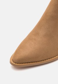 Botín de ante color marrón claro con punta afilada y detalles de costura, que cuenta con una suela de goma flexible para mayor agarre y comodidad.