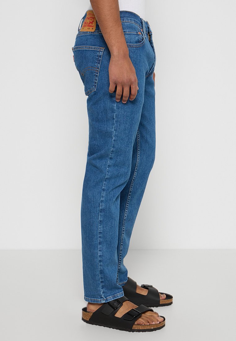 Jeans in denim blu con un taglio dritto, caratterizzati da una patch con logo sulla cintura posteriore, indossati con sandali neri e una camicia bianca.