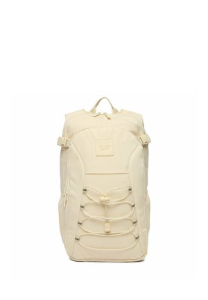 Beżowy plecak Reebok z przednią kieszenią na suwak, regulowanymi bocznymi paskami i elastycznymi sznurkami na przednim panelu.