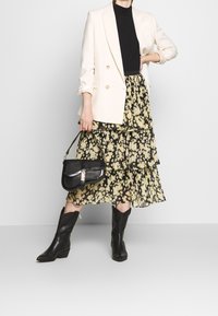 Una mujer se muestra segura de sí misma con un blazer crema sobre una falda floral de volantes, un top negro y botas altas hasta la rodilla también negras, mientras sostiene un pequeño bolso.