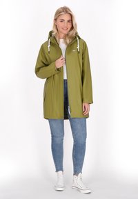 Tamsiai žalia, vandeniui atspari striukė su gobtuvu, spaudimo mygtukais ir raištelių akcentais, dėvima ant balto marškinėlio ir mėlynų džinsų.